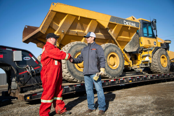 Dusty and TD driver shaking hands with a large dump truck on a trailer in the background.
